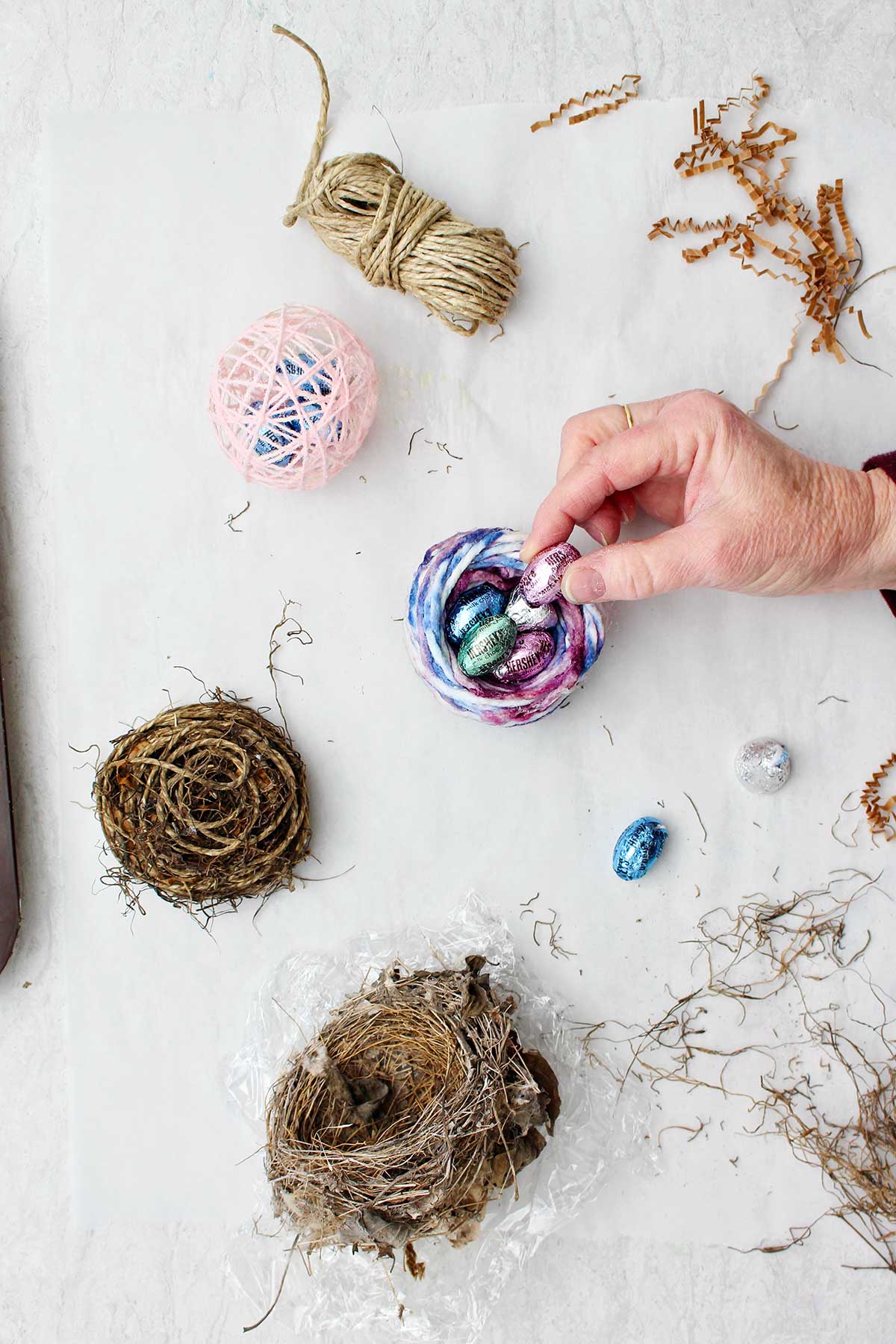 Hand placing Hershey chocolate eggs inside bird nest made of colorful yarn.