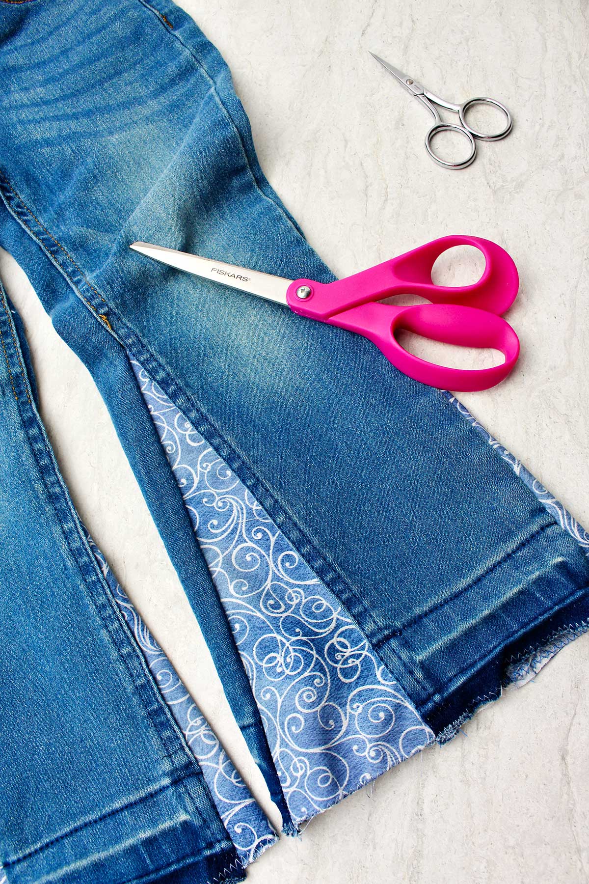 Pair of jeans right side out with decorative fabric sewn on the side to make bell bottoms with two pairs of scissors near by.