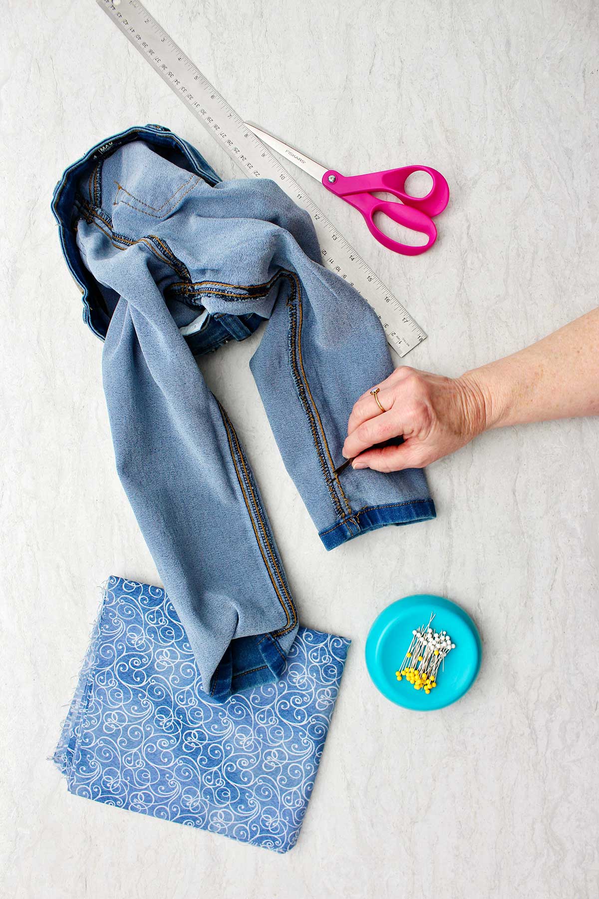 A pair of inside out jeans with hand showing where they want to cut to make bell bottoms with pins, scissors and fabric near by.