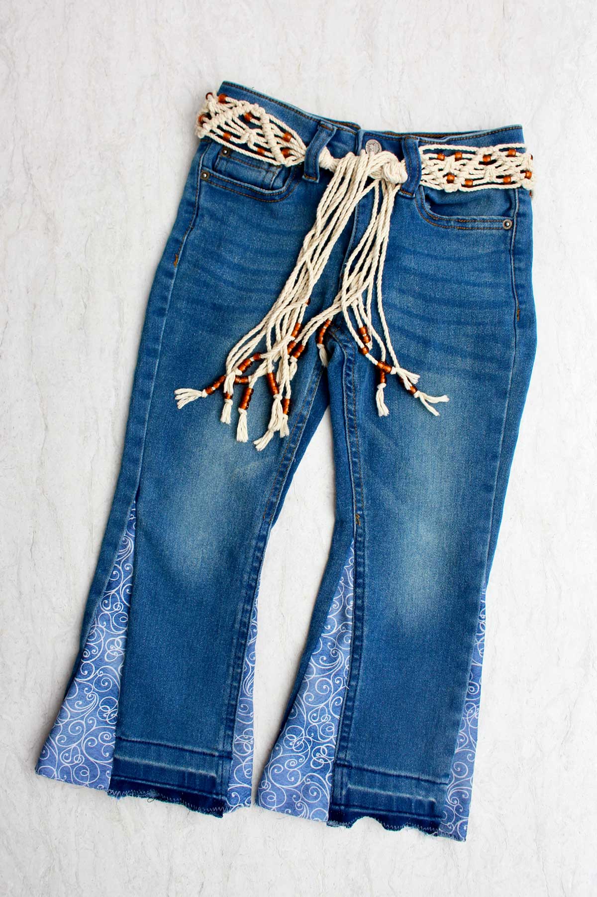 A pair of child's jeans made into bell bottoms with blue swirled fabric, tied with a macrame belt.