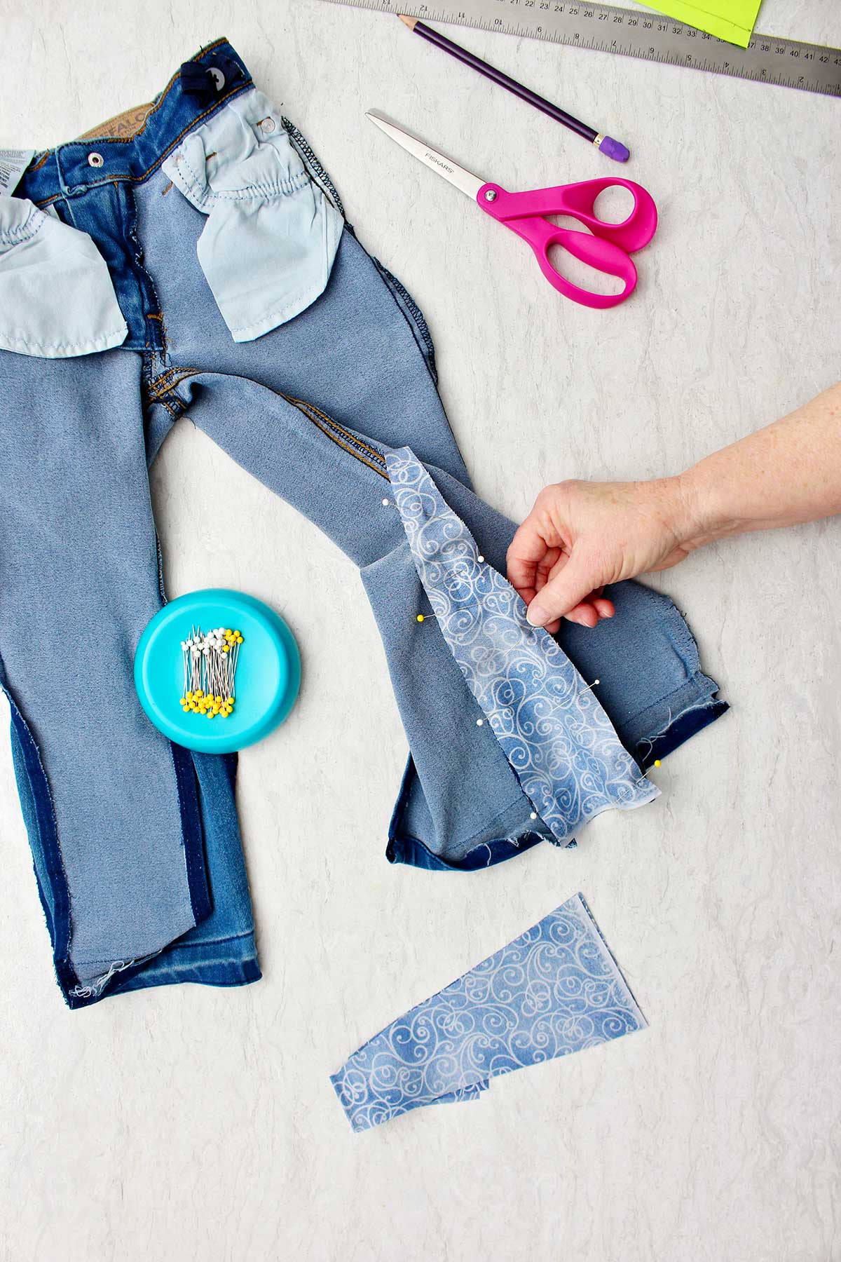 Hand holding fabric secured to jeans with straight pins with supplies near by.