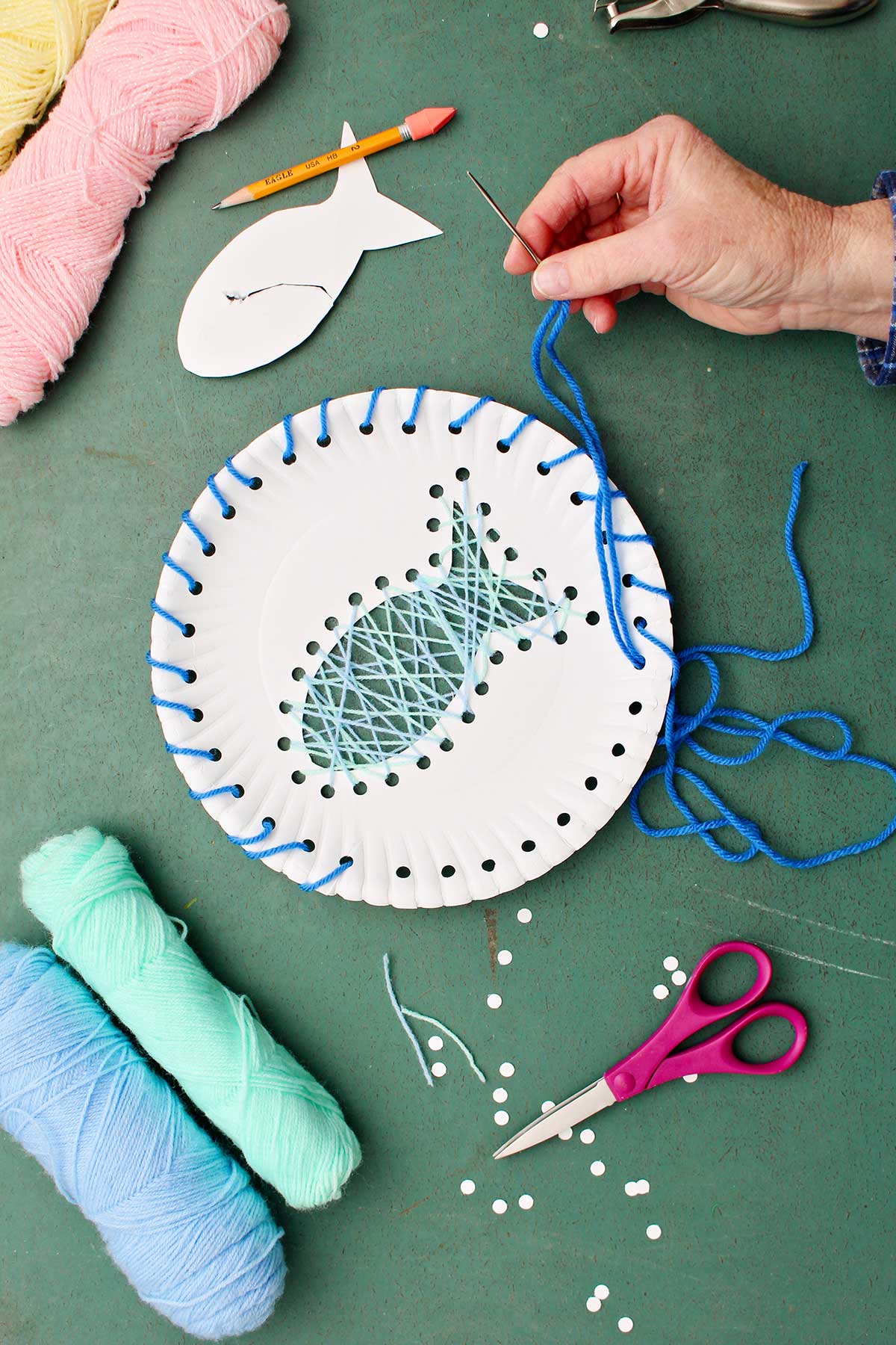Hand threading blue yarn through holes on the outside of paper plate with other supplies near by.