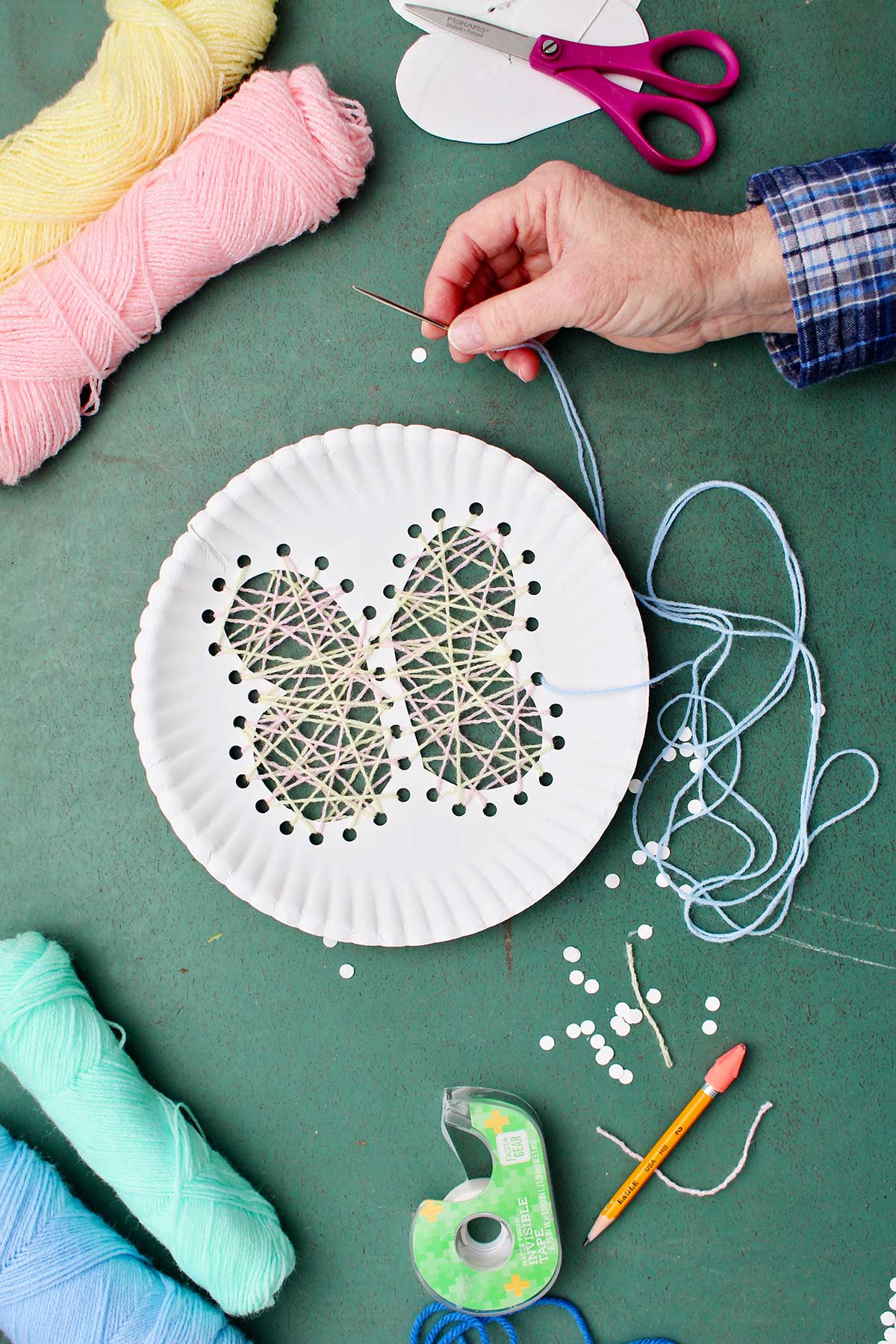 Hand using darning needle and yard to thread through butterfly cut out to make string art project with other supplies near by.