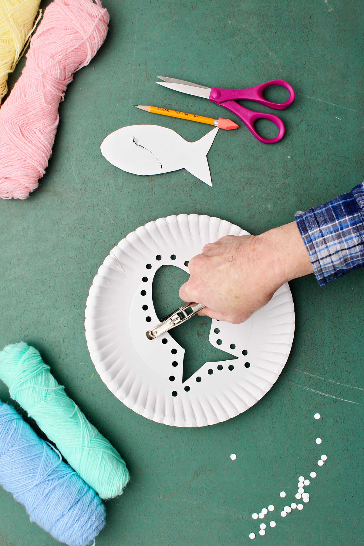 Hand punching holes around fish cut out from paper plate with other supplies near by.
