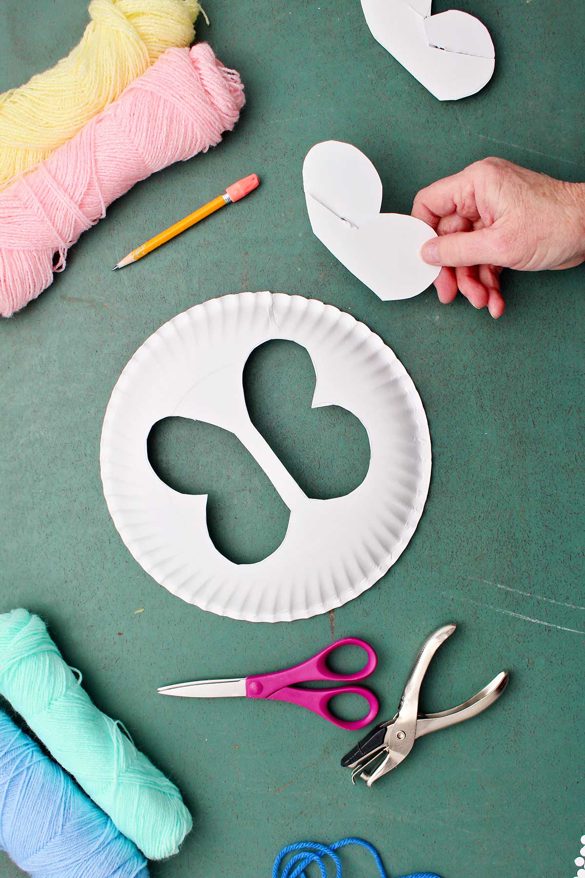 Hand holding butterfly wing cut out from paper plate with yarn and other supplies near by.