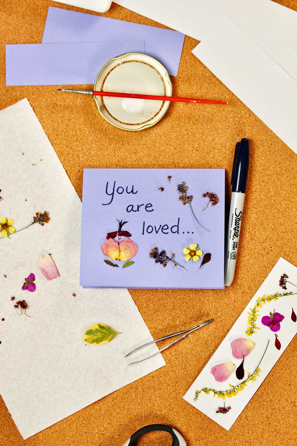 A purple card saying "You are loved..." with pressed flowers on it as well as a bookmark with pressed flowers.