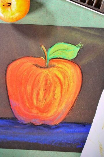 Drawing of apple, fresh apple and pastels on a green counter.