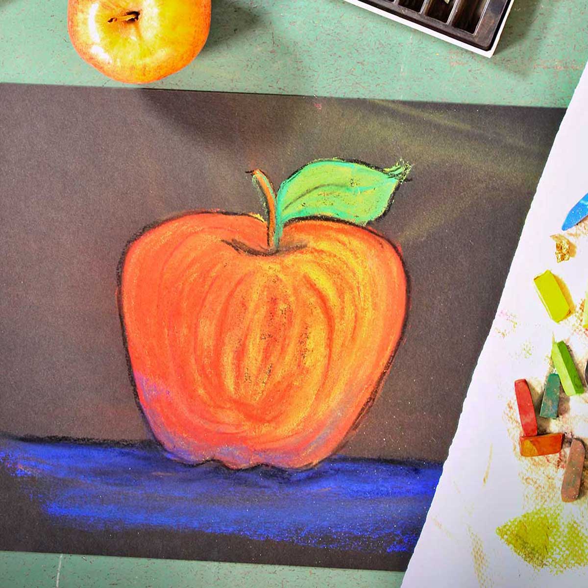 Drawing of apple, fresh apple and pastels on a green counter.