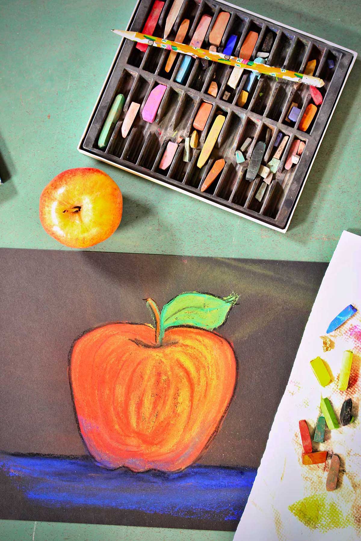Drawing of apple, fresh apple and pastels on a green counter.