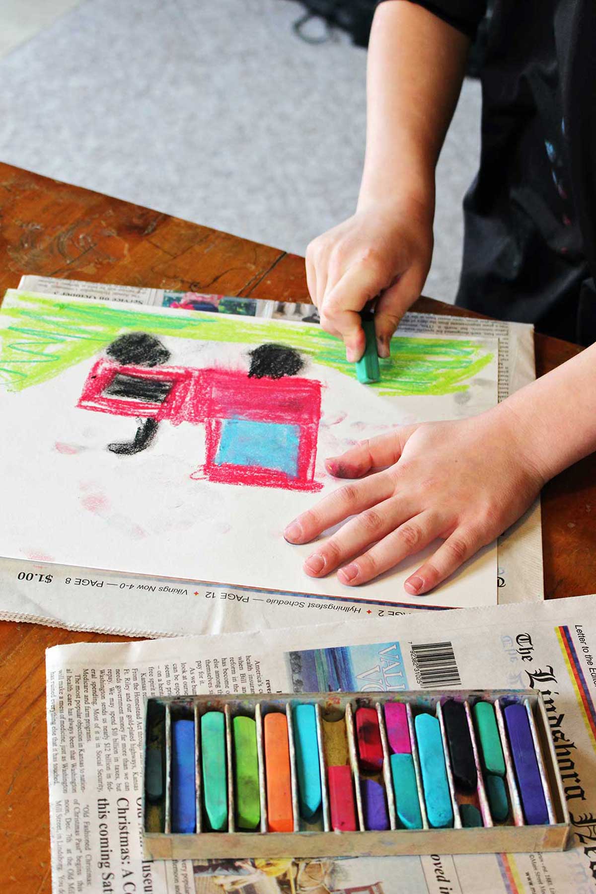 Child coloring a picture of a red tractor on green grass with pastels.