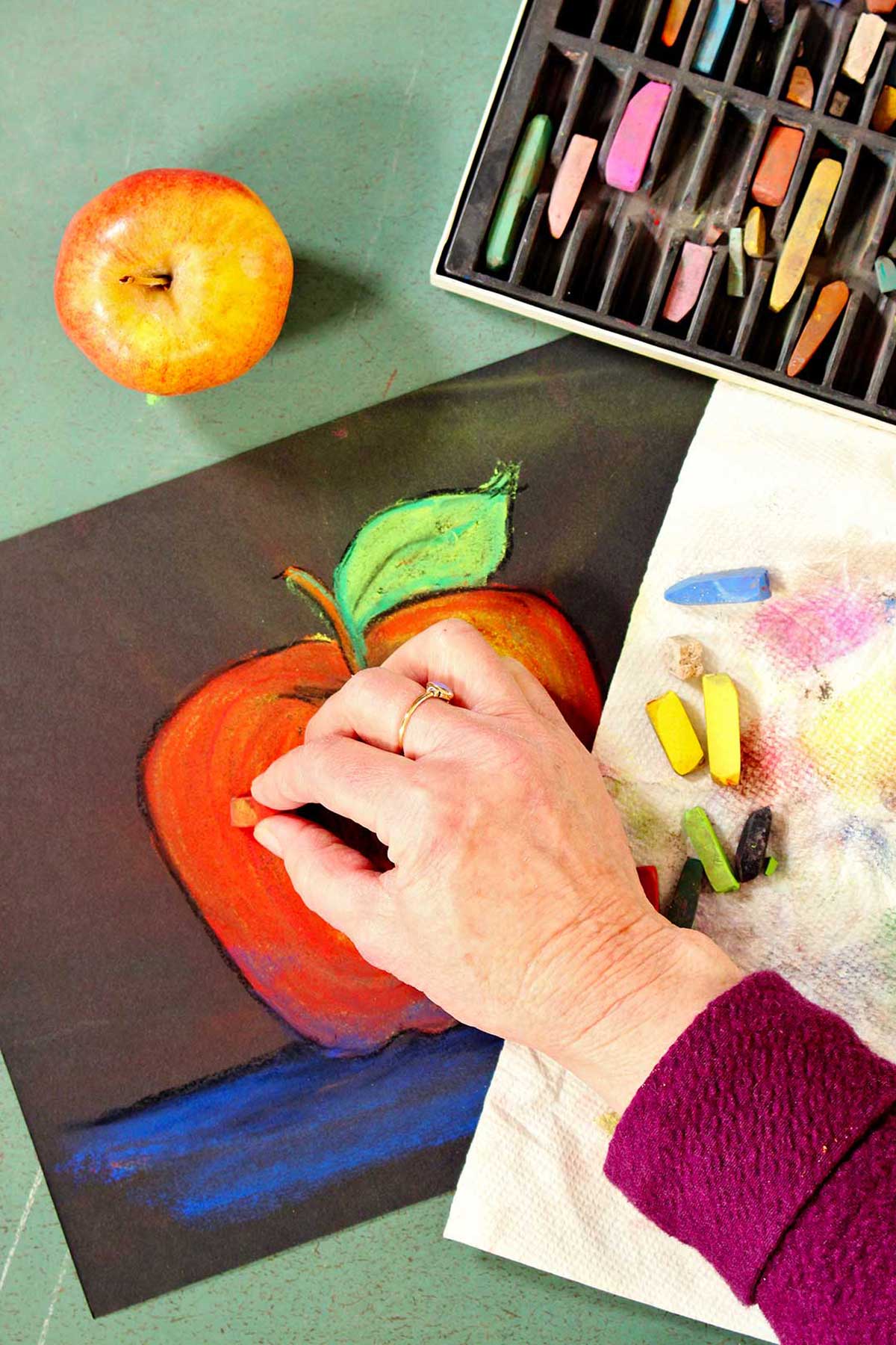 Hand drawing red apple on a black piece of construction paper with pastels and a red apple is near by.