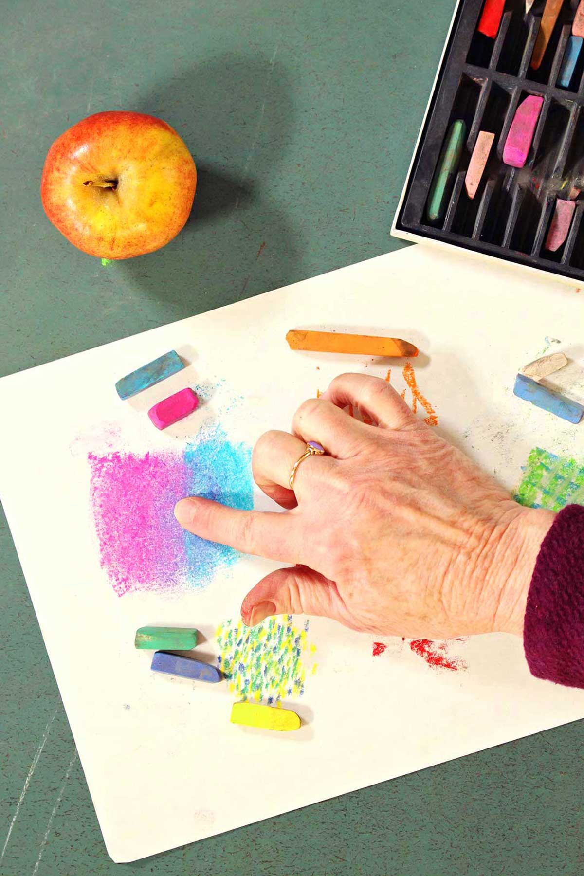Hand blending colors of pastels on a piece of paper with pastels and a fresh apple on green counter.