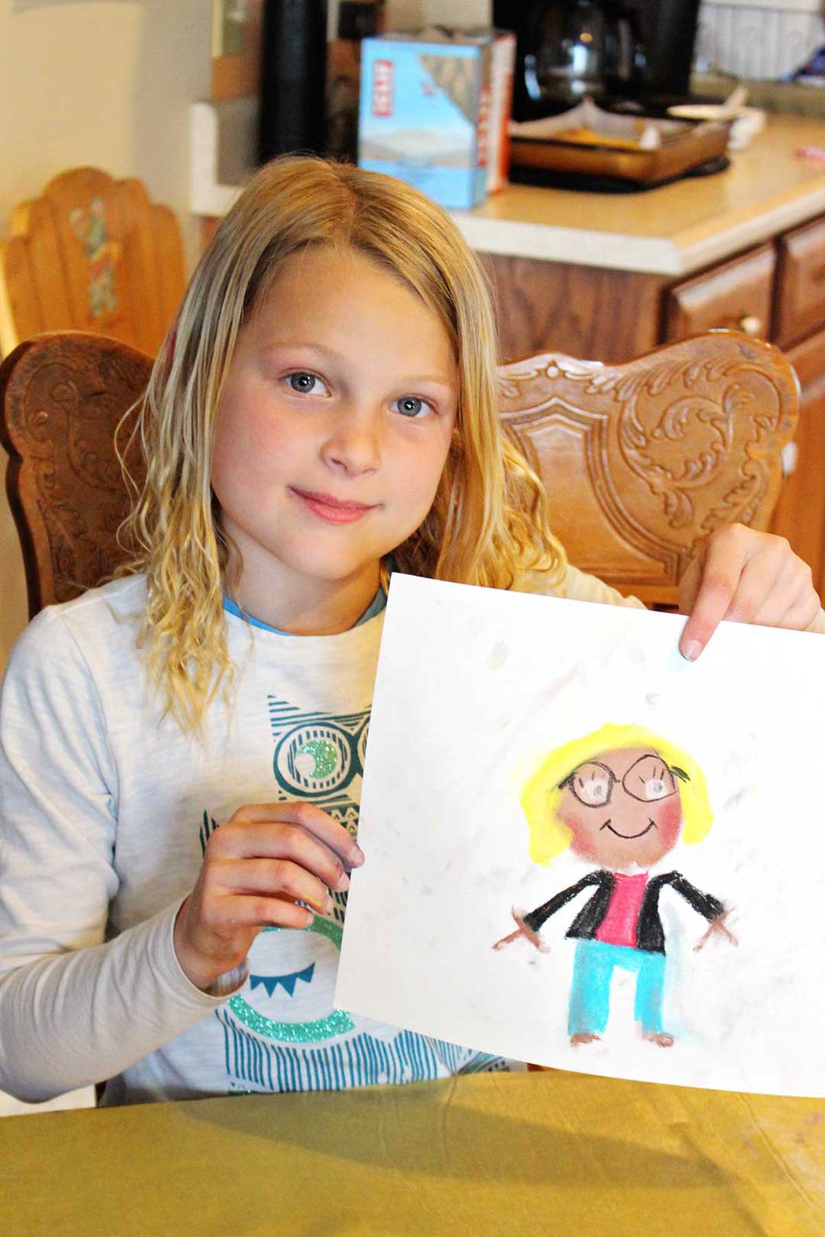 Young girl with blonde hair sits at a kitchen table holding up her drawing of a girl with blonde hair and glasses with blue pants and a black blazer.