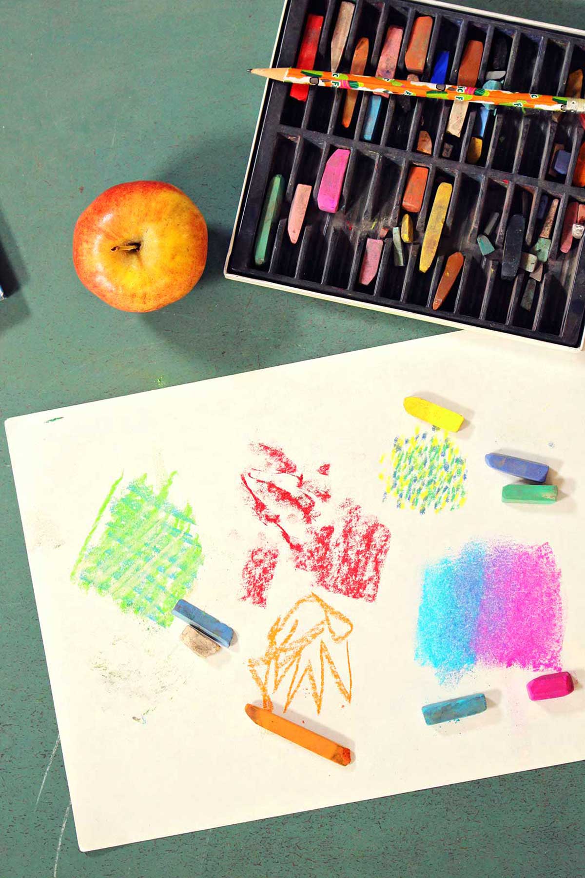 Doodles on a white piece of paper using pastels with tray of pastels and apple next to it.