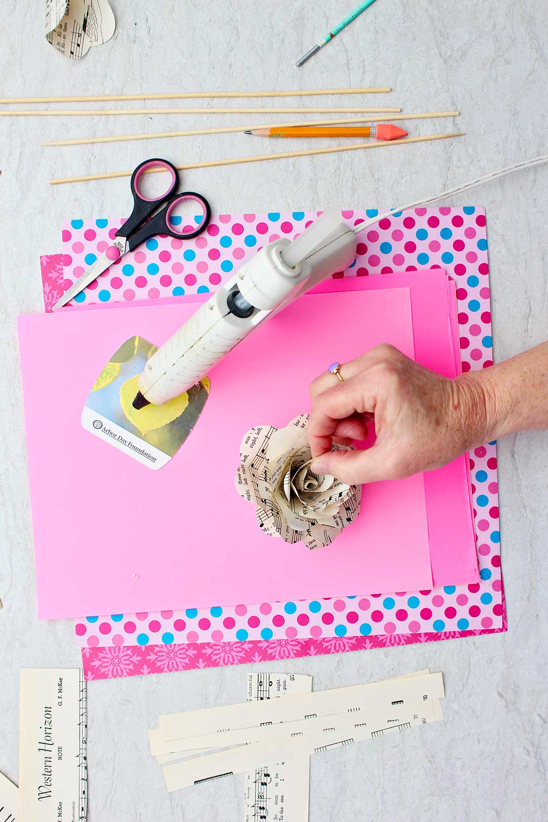 Hand placing paper in the center of an almost completed paper rose sitting on a stack of pink scrapbook paper with other supplies near by.