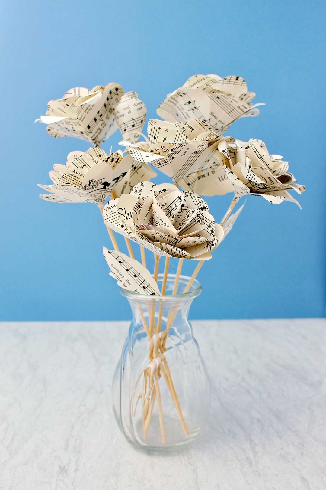 Completed Book Page Roses made from sheet music on wooden dowel stems in a glass vase with a blue background behind it.