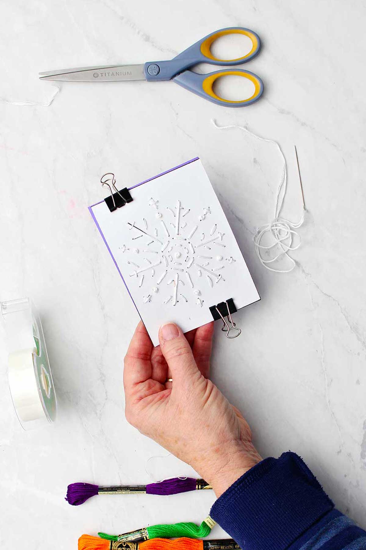 Hand holding embroidered snowflake paper clamped on purple card with embroidery floss, tape and scissors nearby.