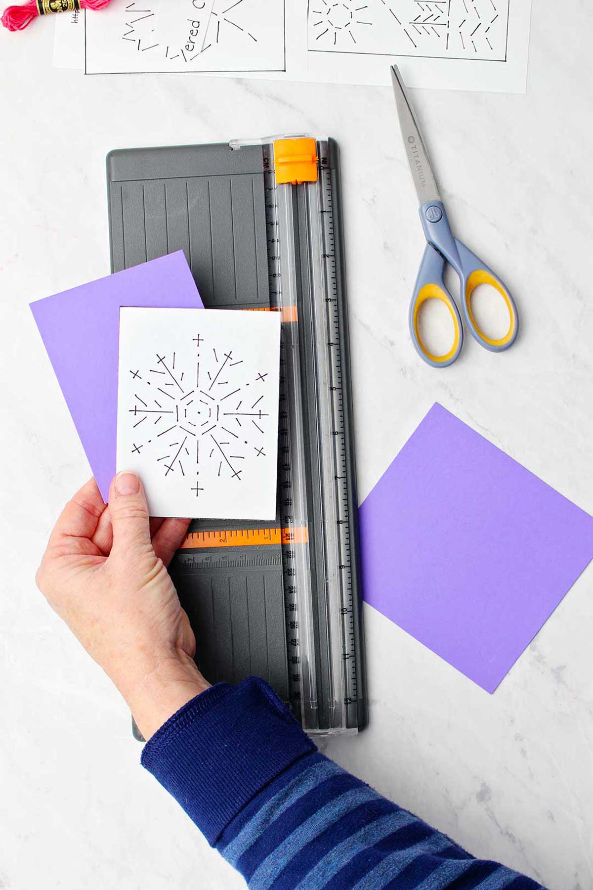 A hand holding card and snowflake template resting on a paper cutter.