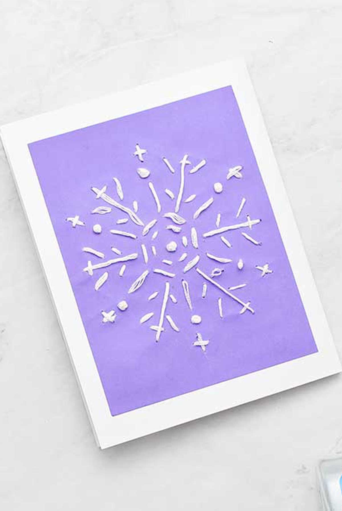 Completed snowflake embroidered greeting card with embroidery floss, glue and scissors nearby.