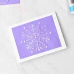 Completed snowflake embroidered greeting card with embroidery floss, glue and scissors nearby.