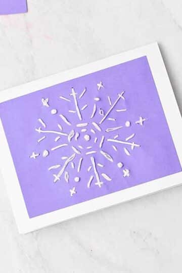 Completed snowflake embroidered greeting card with embroidery floss, glue and scissors nearby.