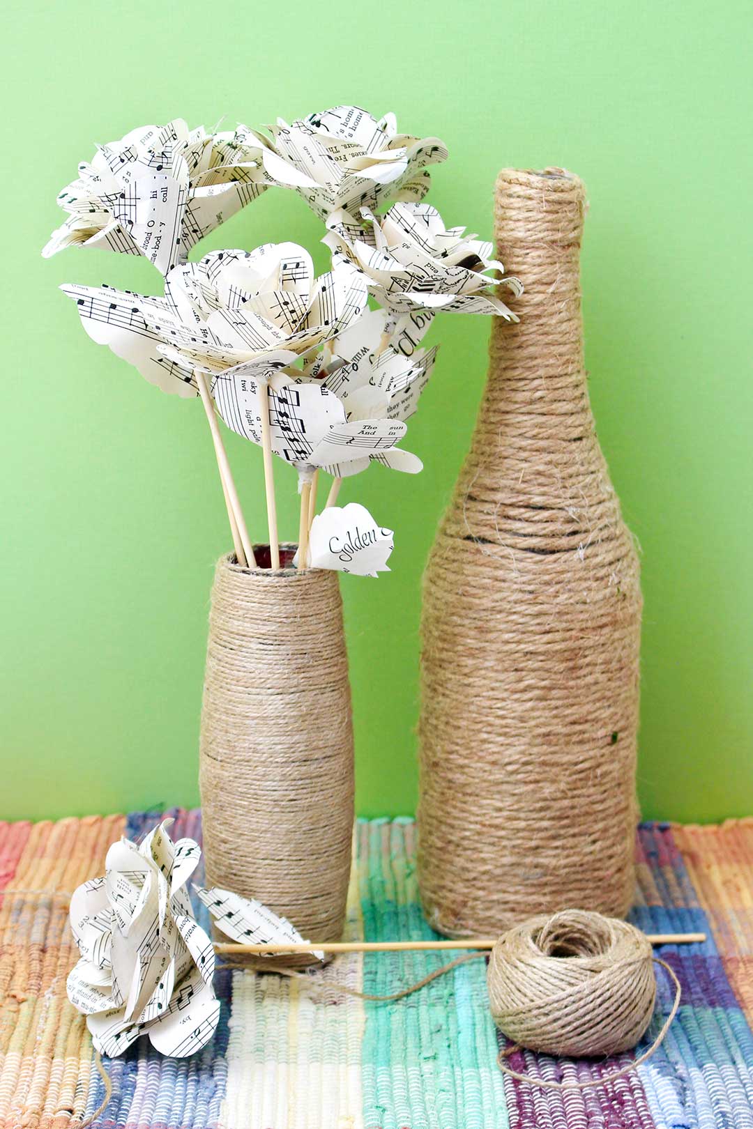 A twine wrapped bottle and a twine wrapped vase with paper flowers inside sitting on a colorful placemat against a green backdrop.