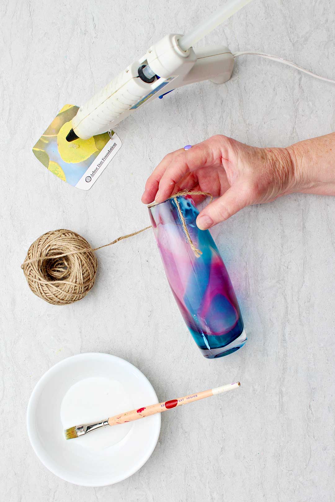 Hand holding glass vase with the starting of the twine wrapping glued to it with glue gun and white glue with paint brush near by.