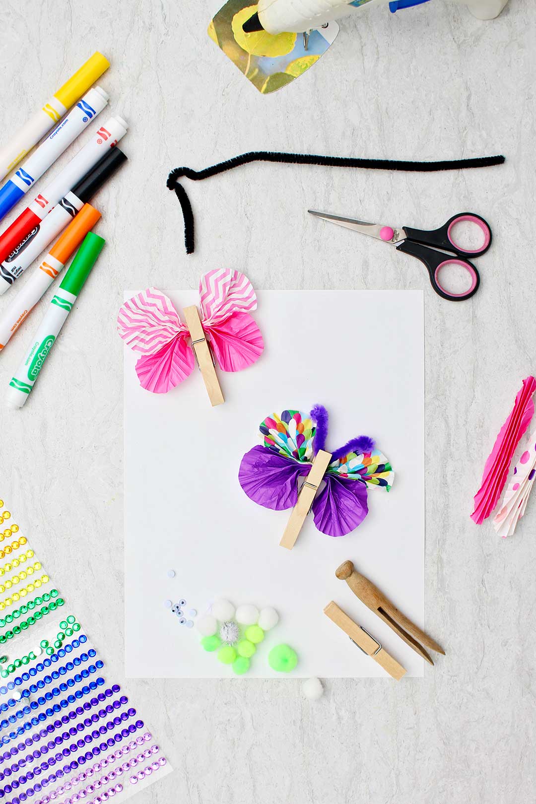 Two wooden clothespins clipping colorful paper wings sitting on a white piece of paper with other crafting supplies near by.