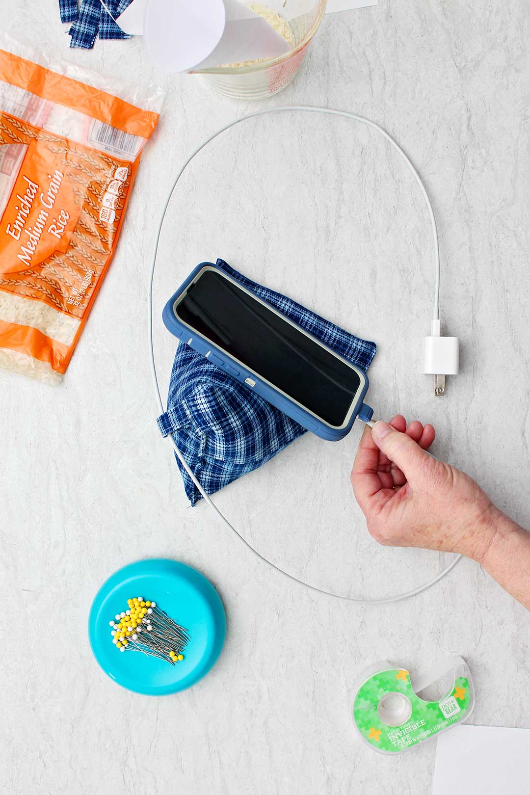 Hand plugging in cell phone charger into cell phone which is resting on completed Cell Phone Holder Pillow with other supplies near by.