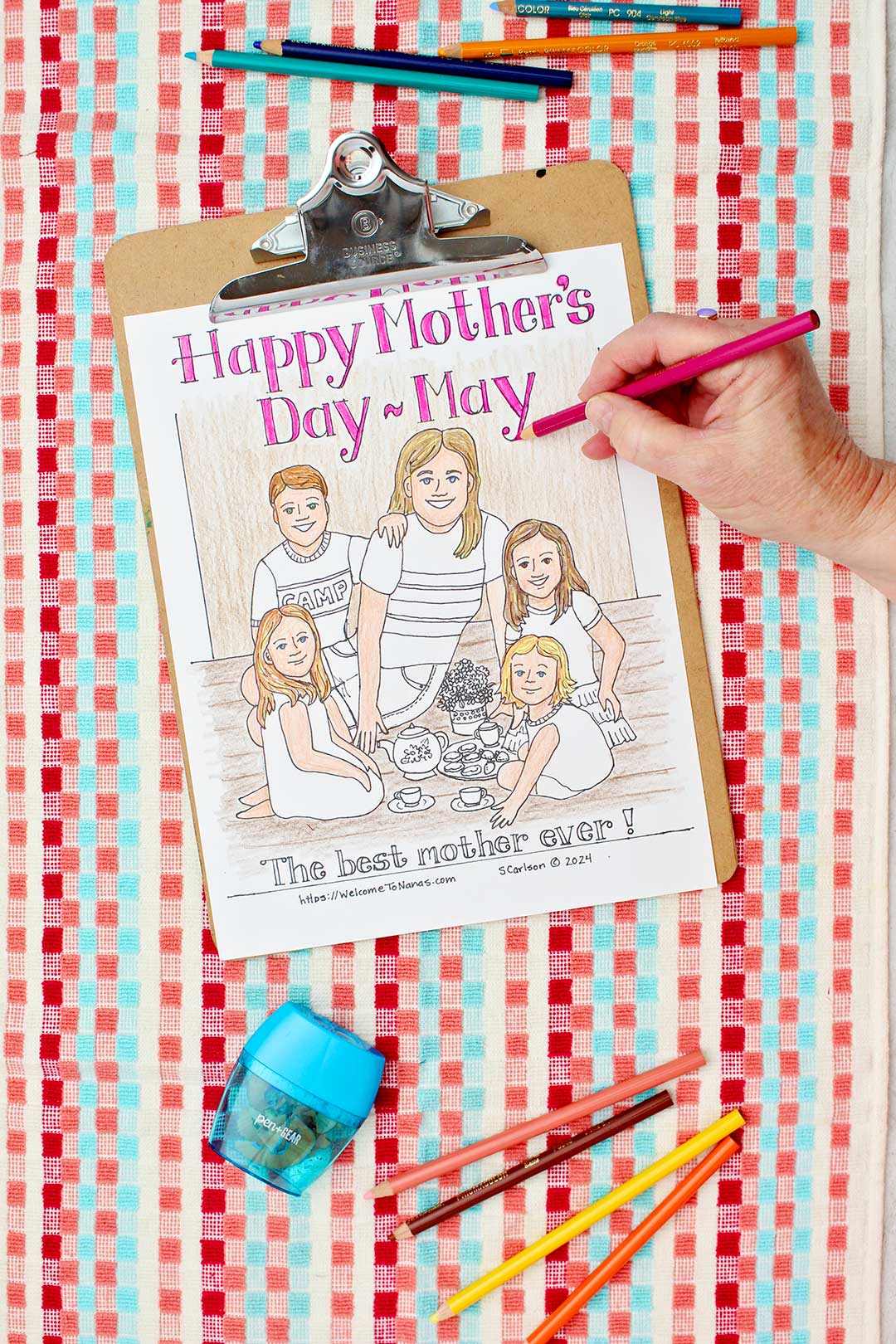 Hand coloring text pink on Mother's Day Coloring page of a mom and kids having a tea party clipped into clip board and resting on a red, coral and aqua towel with colored pencils near by.