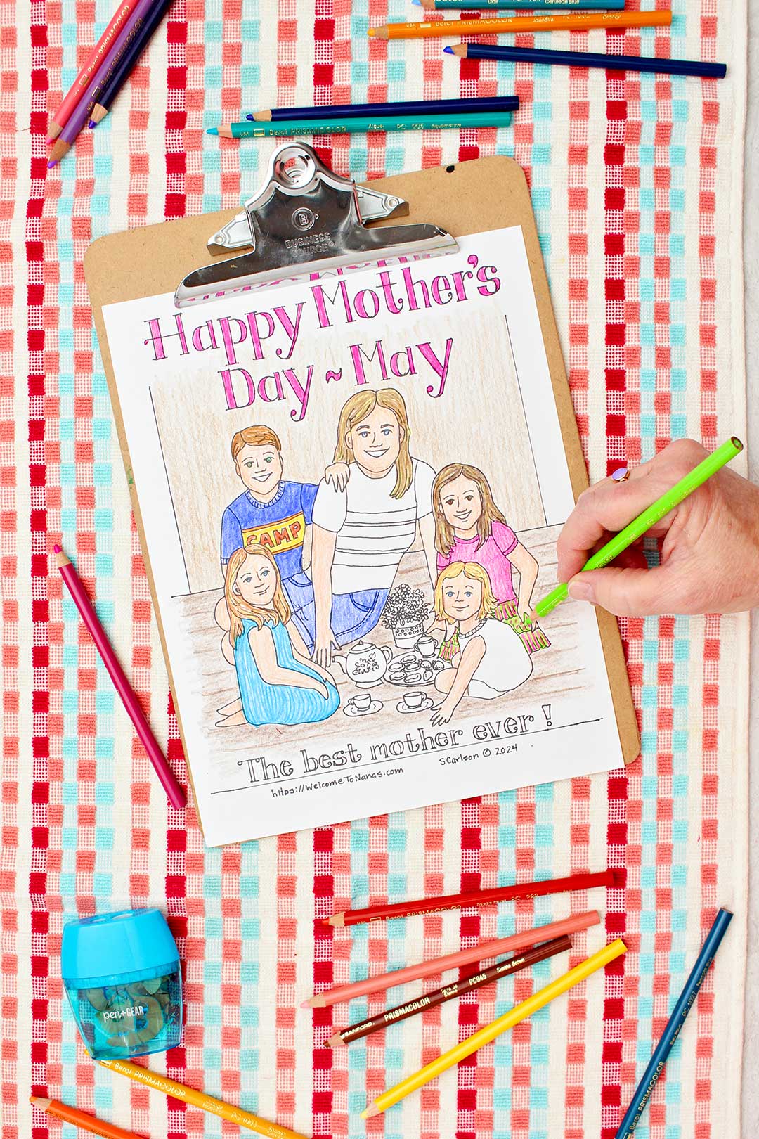 Hand coloring girl's dress green on Mother's Day Coloring page of a mom and kids having a tea party clipped into clip board and resting on a red, coral and aqua towel with colored pencils near by.
