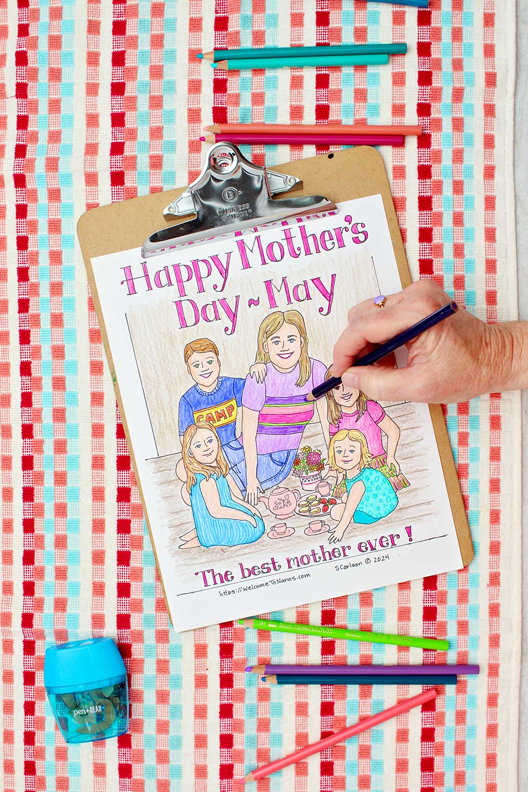 Hand coloring stripes on mom's shirt purple on Mother's Day Coloring page of a mom and kids having a tea party clipped into clip board and resting on a red, coral and aqua towel with colored pencils near by.