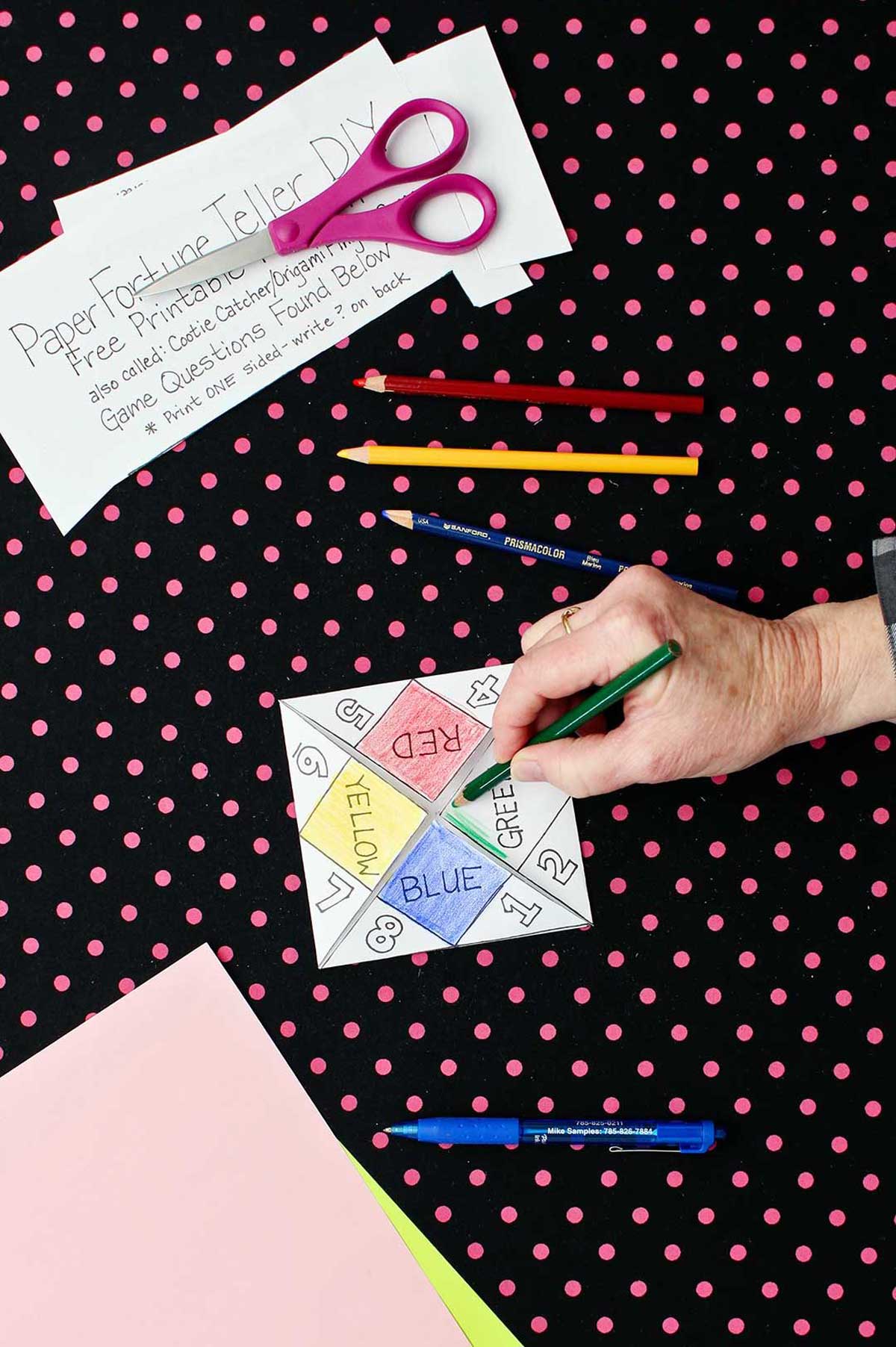 Hand coloring green tab green on printout of paper fortune teller with paper, scissors and colored pencils resting on black and pink polka dotted background.