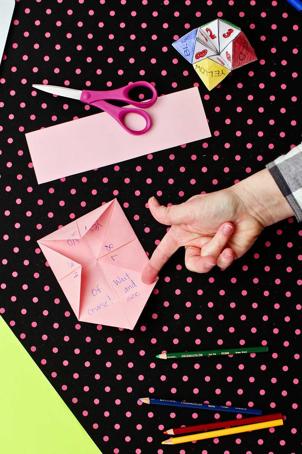 Inside flaps of the pink paper fortune teller stating fortunes with scissors and colored pencils laying on a black and pink polka dotted background.
