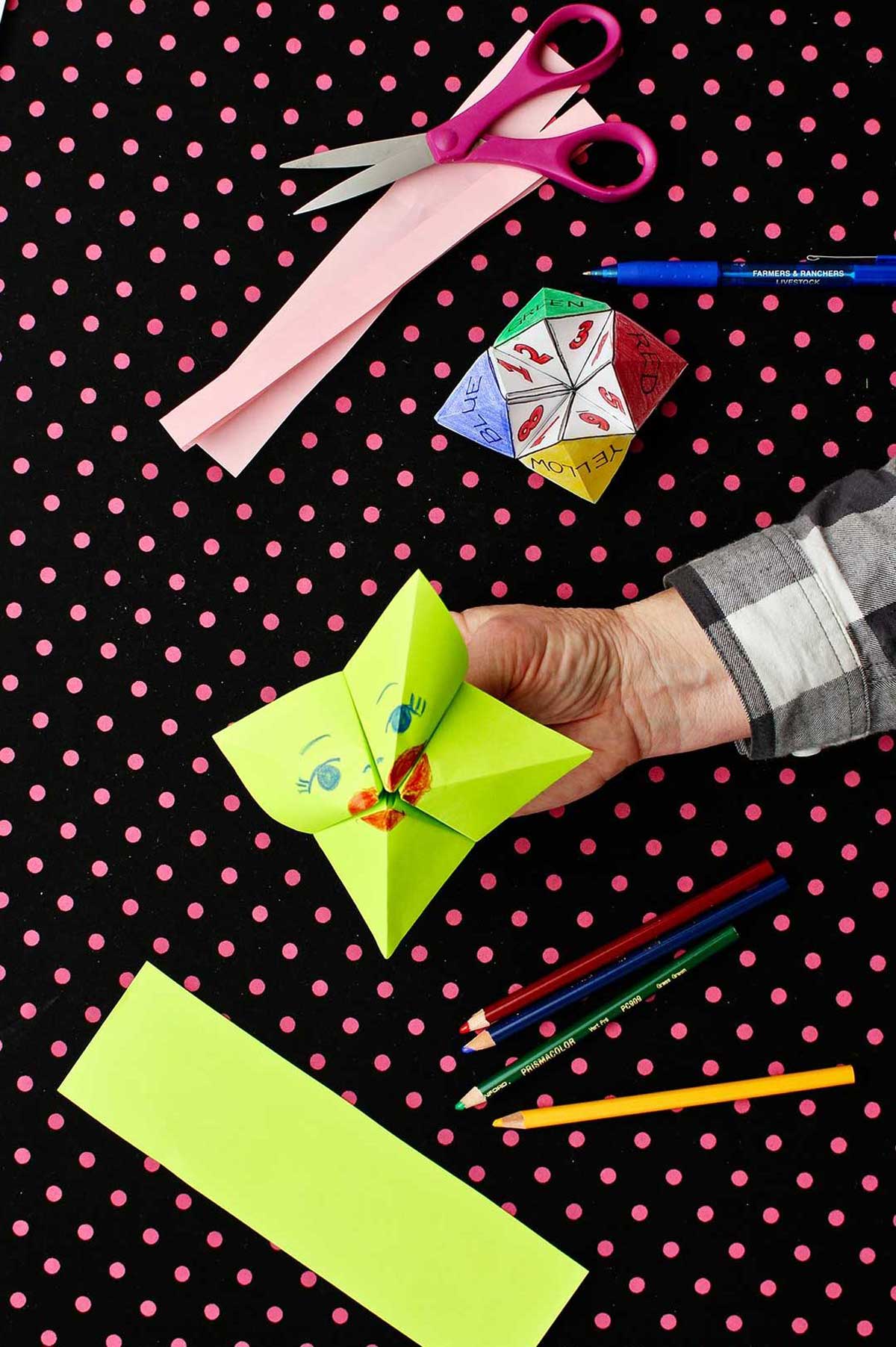Hand holding green completed fortune teller with a face on the front with various supplies near by against black and pink polka dotted background.