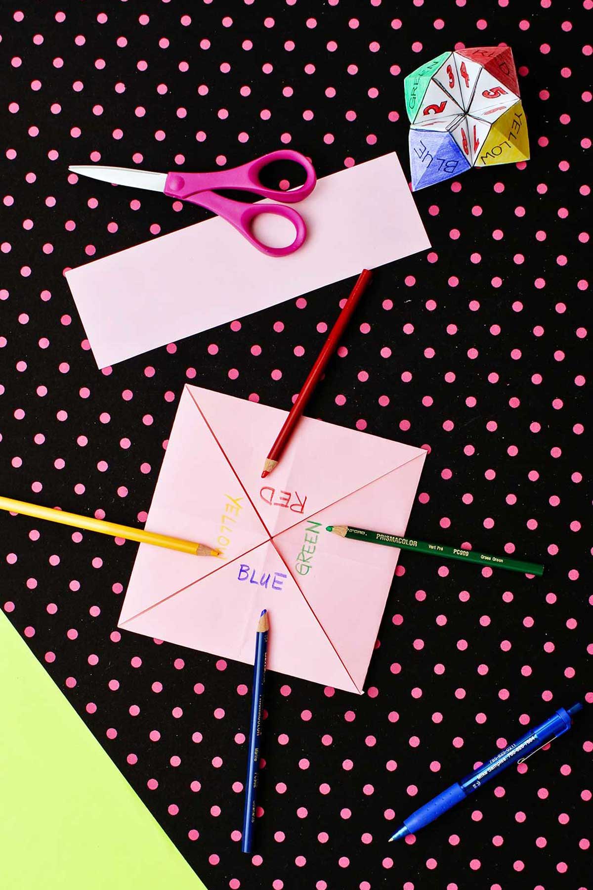 Four tabs on a square pink piece of paper saying "red, green, yellow, blue" with scissors and colored pencils laying on a black and pink polka dotted background.