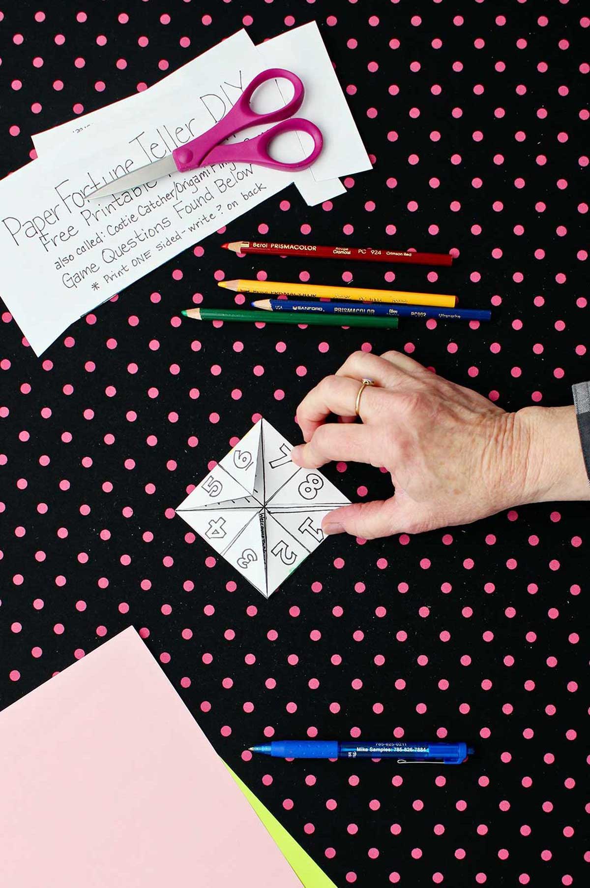 Hand showing folded flaps and numbers on printout of paper fortune teller with paper, scissors and colored pencils resting on black and pink polka dotted background.