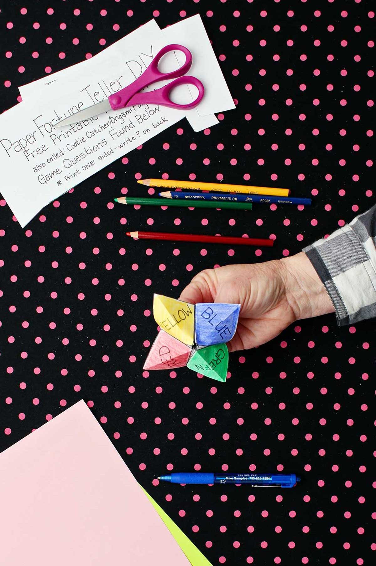 Hand holding completed and colored printout of paper fortune teller with paper, scissors and colored pencils resting on black and pink polka dotted background.