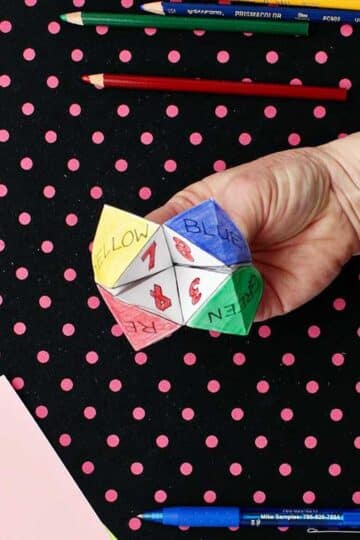 Hand opening completed and colored printout of paper fortune teller with paper, scissors and colored pencils resting on black and pink polka dotted background.