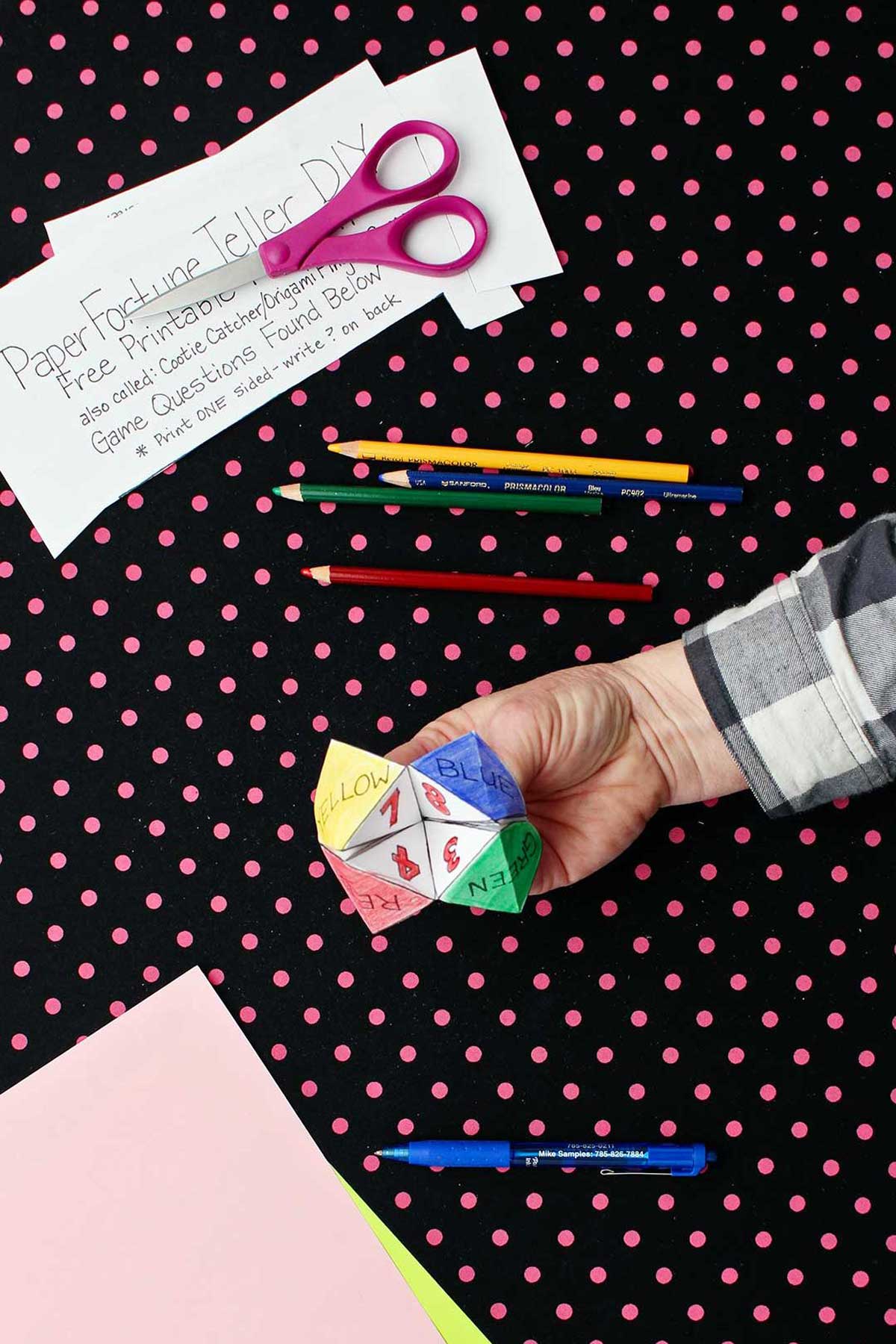 Hand opening completed and colored printout of paper fortune teller with paper, scissors and colored pencils resting on black and pink polka dotted background.