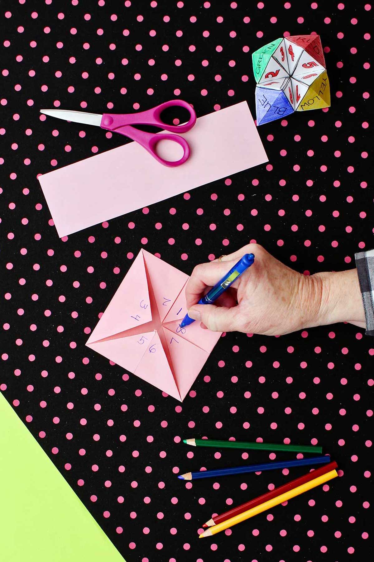 Opposite side of paper fortune teller made from pink paper showing numbers 1-8 with scissors and colored pencils laying on a black and pink polka dotted background.