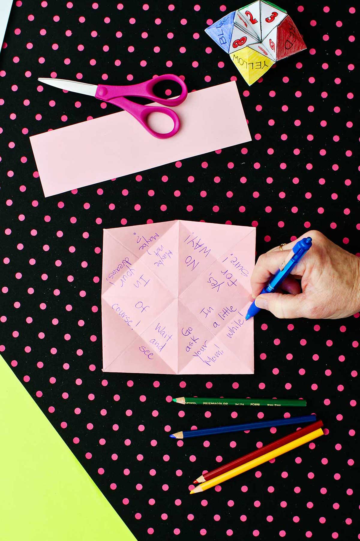 Unfolded square pink paper with all the options for fortunes for the paper fortune teller with scissors and colored pencils laying on a black and pink polka dotted background.