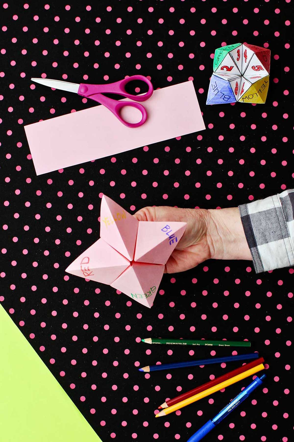 Hand holding completed paper fortune teller made of pink paper with "red, yellow, green, blue" tabs with scissors and colored pencils laying on a black and pink polka dotted background.