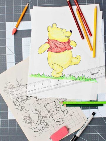 A completed, enlarged and colored picture of Winnie the Pooh, colored pencils, ruler, eraser, Sharpie, and original Pooh image with grid drawn over it resting on a drawing mat.