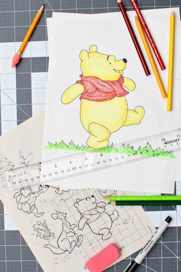 A completed, enlarged and colored picture of Winnie the Pooh, colored pencils, ruler, eraser, Sharpie, and original Pooh image with grid drawn over it resting on a drawing mat.