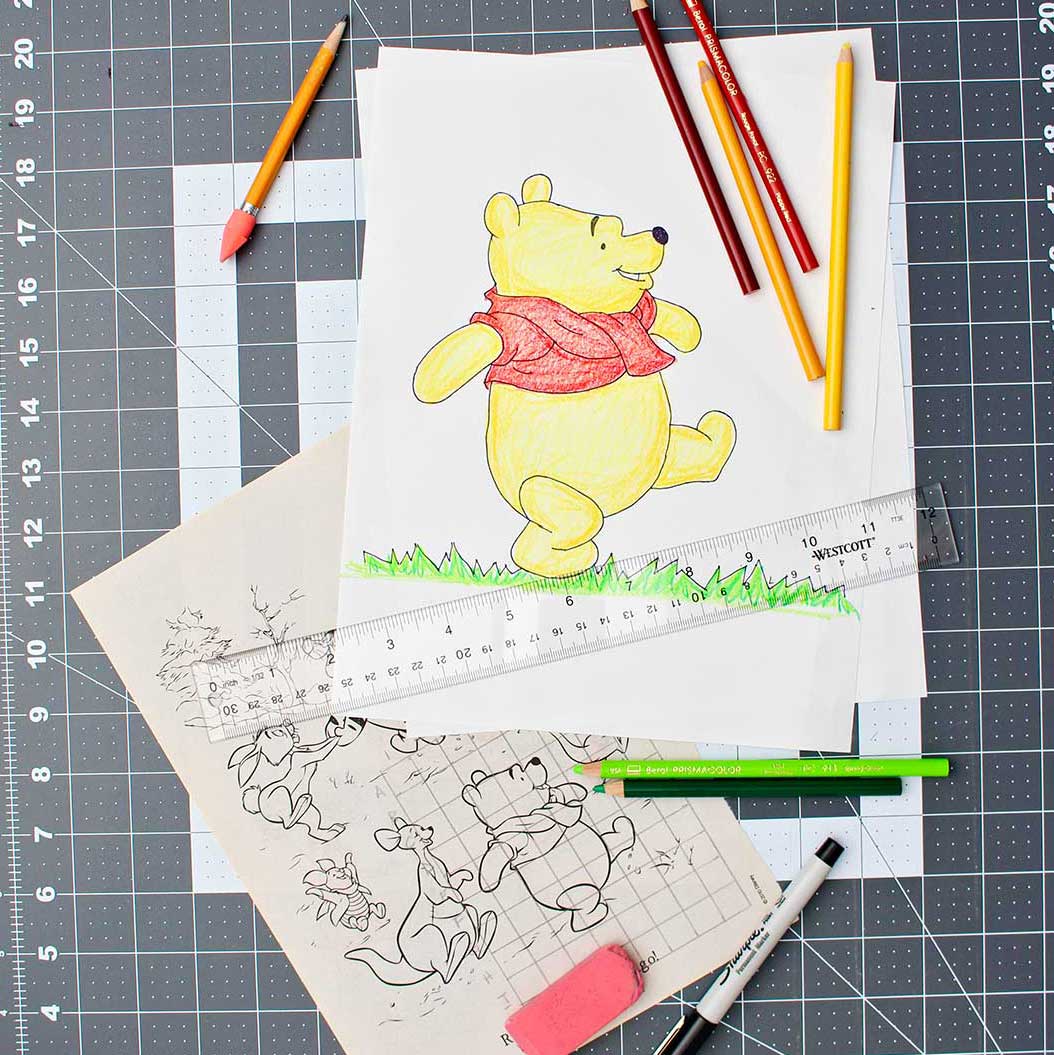 A completed, enlarged and colored picture of Winnie the Pooh, colored pencils, ruler, eraser, Sharpie, and original Pooh image with grid drawn over it resting on a drawing mat.