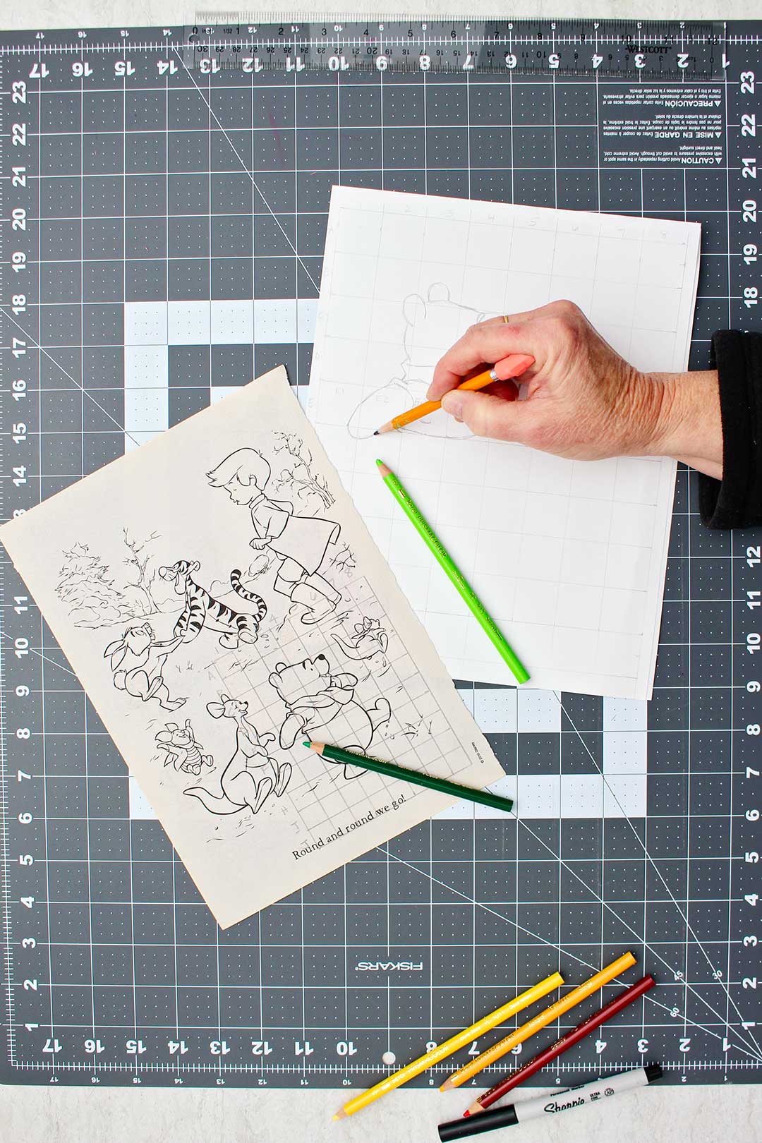 Hand sketching arm of Winnie the Pooh in the enlarged sketch with the original image near by.