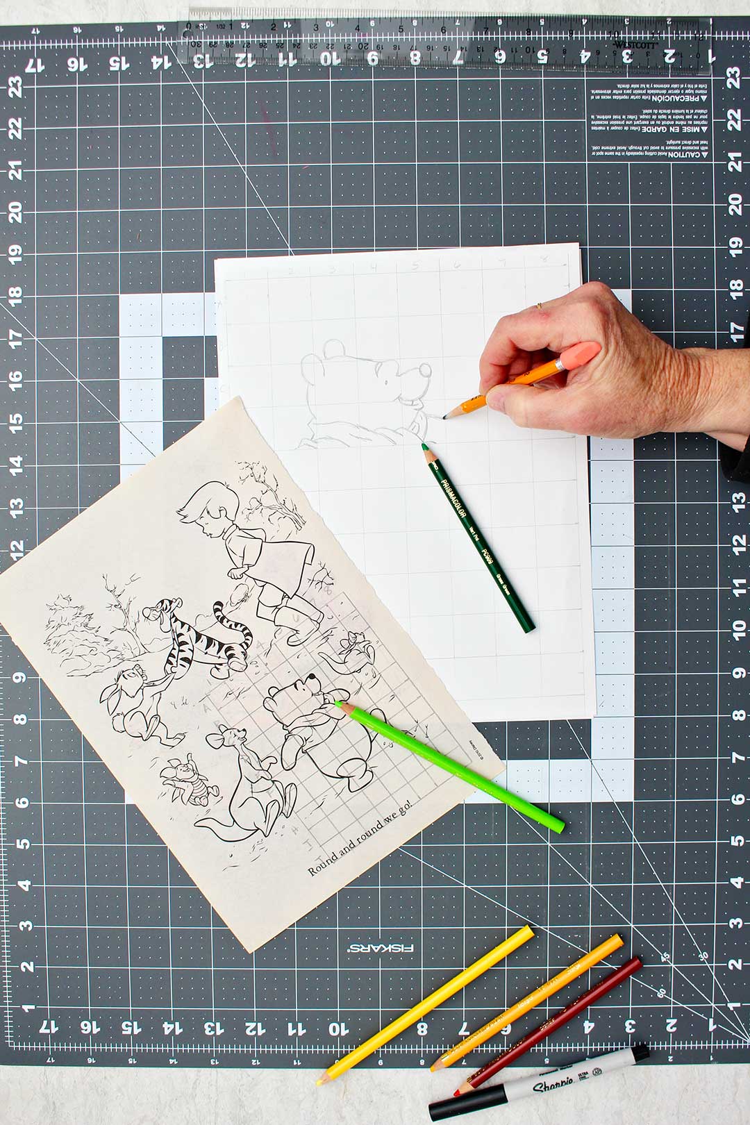 Hand sketching arm of Winnie the Pooh in the enlarged sketch with the original image near by.