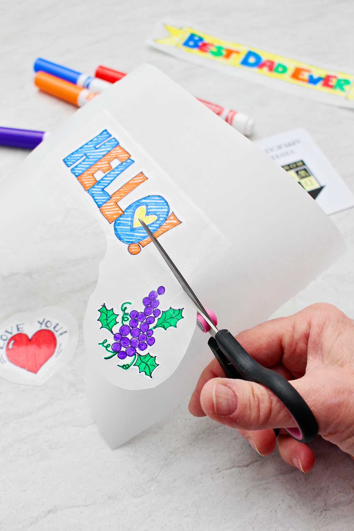 Hand cutting out stickers from parchment tape sheet. A bunch of grapes and a sticker that says "HELLO" in block letters with other supplies in the background.