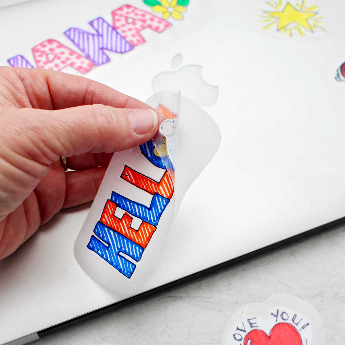 Hand peeling back a sticker that says "HELLO" with silver laptop and stickers in the background.